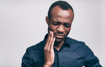 Feeling toothache. Frustrated young African man touching his cheek and keeping eyes closed while standing against grey background